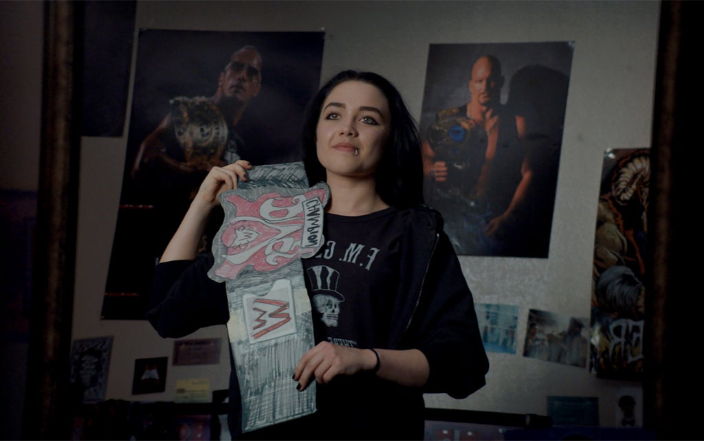 A person with dark hair and a nose ring, wearing a black sweatshirt with a graphic design, holds up a hand drawn wrestling belt . Behind them, the wall is decorated with several posters, including images of famous wrestlers presenting their championship belts in similar fashion