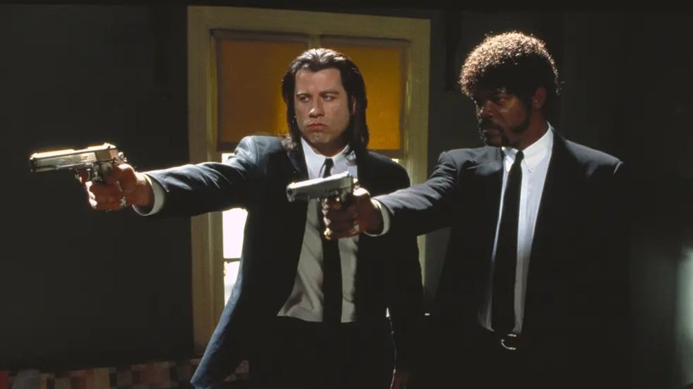 Two men in dark suits and ties stand side by side in a dimly lit room, each pointing a handgun with serious expressions. The man on the left has long dark hair, while the man on the right has a short afro and goatee. Behind them, sunlight streams through a window with orange blinds, casting dramatic shadows in the room.
