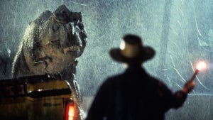 A large dinosaur stands in the foreground, its head lowered and mouth open, revealing sharp teeth. Behind the dinosaur, a person wearing a cowboy hat holds a flare, illuminating the dark, rainy scene. The background features a blurred vehicle with headlights on, and water droplets are visible in the air.