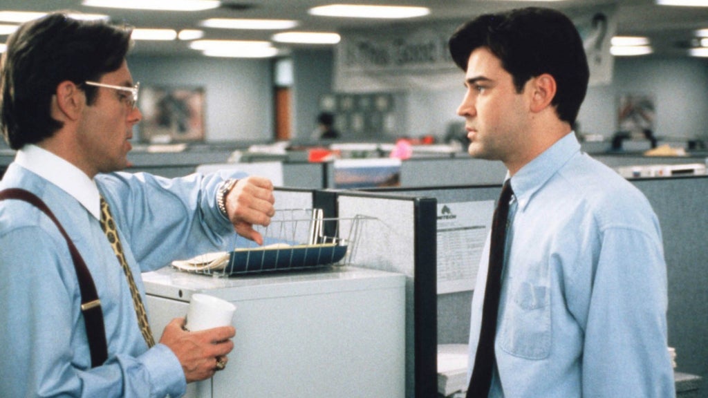Two men stand facing each other in an office setting, separated by a cubicle wall. The man on the left, wearing glasses, a light blue shirt, and suspenders, holds a white coffee cup and extends his other hand toward a basket on the cubicle wall. The man on the right, dressed in a light blue shirt and tie, looks at the basket with a neutral expression. Behind them, the office environment includes desks, computers, and overhead lighting.