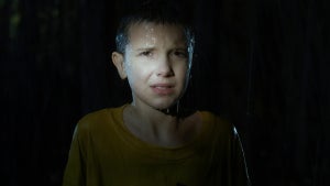 A young girl stands drenched in heavy rain, illuminated by a harsh spotlight that highlights her distressed expression. The surrounding darkness makes her look vulnerable and alone.