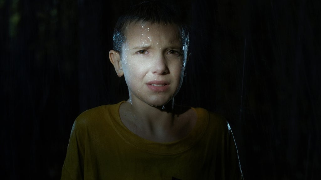 A young girl stands drenched in heavy rain, illuminated by a harsh spotlight that highlights her distressed expression. The surrounding darkness makes her look vulnerable and alone.