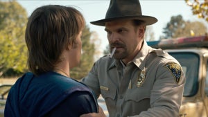 A police officer in a tan uniform grabs a teenage boy by the shoulders, speaking to him intensely. They stand outside near a patrol car in broad daylight.