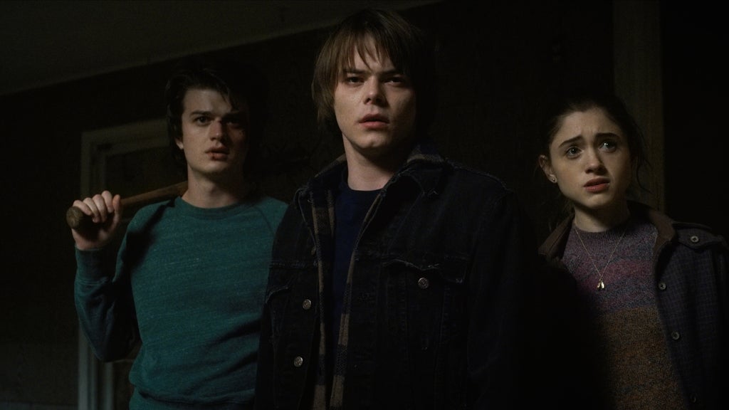 Three teenagers stand in a dark, tense hallway, illuminated by dim light. Steve grips a nail-studded baseball bat, Jonathan looks shocked and unsteady, and Nancy stares ahead with wide, worried eyes. All three appear braced for danger.