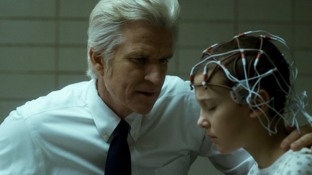 A stern-looking scientist leans close to a young girl seated in a clinical lab setting. The girl wears a medical gown and an array of wires and electrodes on her shaved head. She looks tense and exhausted as he whispers something to her.