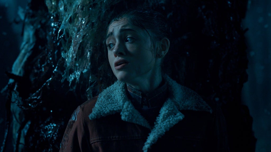 A teenage girl stands in a dark, eerie environment covered in wet, organic-looking vines. She looks over her shoulder with fear and urgency as blue-tinted light reveals the slimy, otherworldly textures around her.