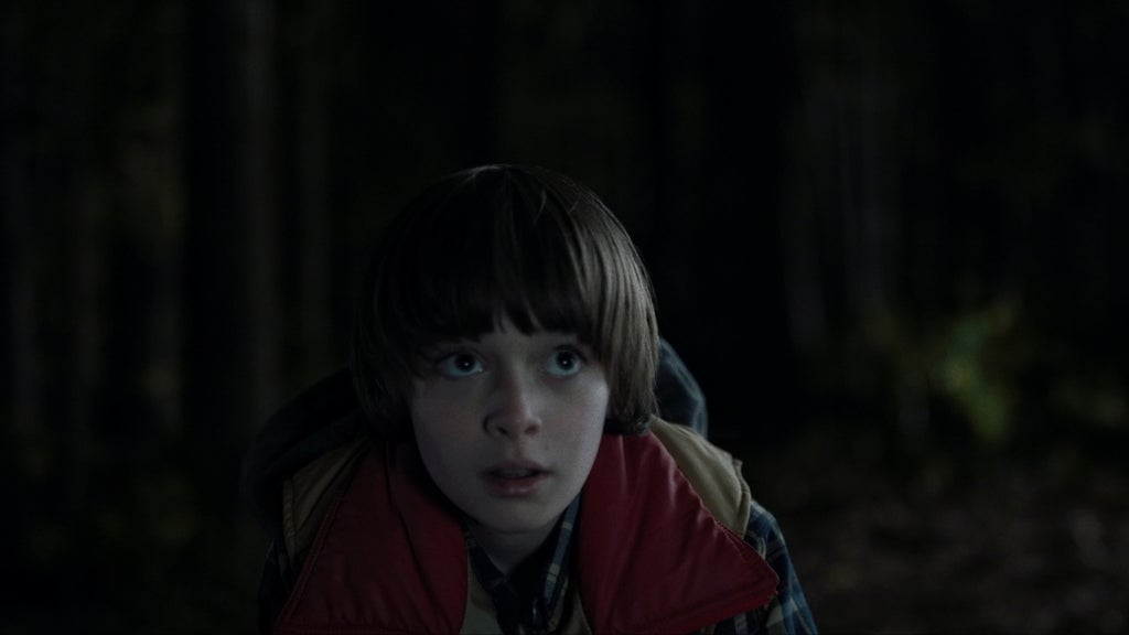 A young boy crouches alone in a dark forest at night, staring upward with frightened, wide eyes. He wears a red vest over a plaid shirt, and the shadows around him create an eerie, isolated atmosphere.