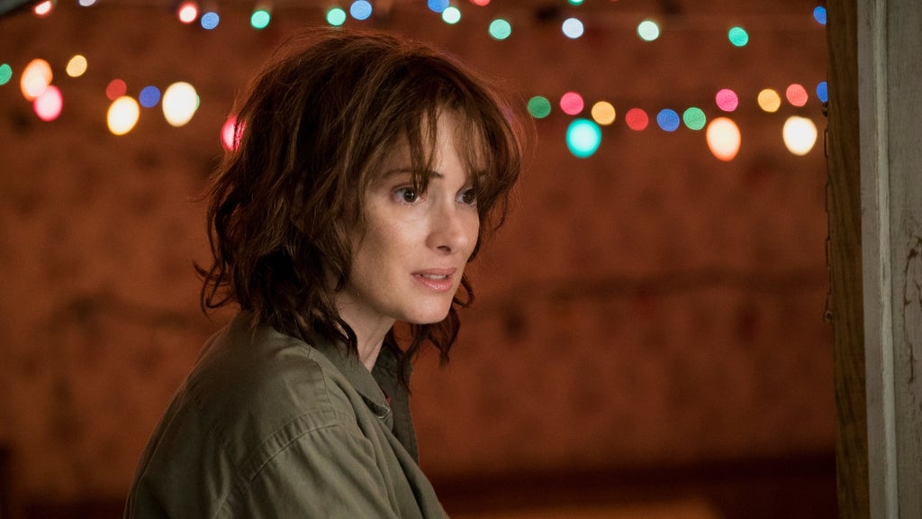 A worried woman stands in a cluttered room illuminated by colorful Christmas lights strung across the wall behind her. Her hair is tousled and she looks tense, as if listening for something unseen.