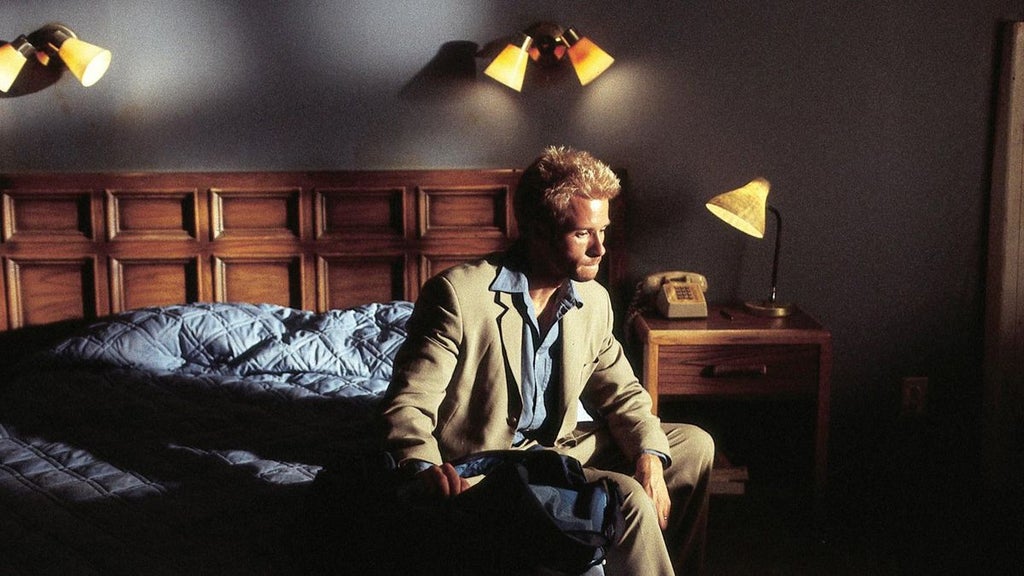 A man with bleached hair sits on the edge of a bed in a dimly lit room with wooden paneling on the walls. He is wearing a beige suit with a blue shirt and tie, holding a small object in their hands. Behind him, the bed has a dark bedspread with a patterned pillow. A wooden nightstand with a lamp and a vintage telephone is beside the bed. Overhead, three wall-mounted lamps with yellow shades provide soft lighting.
