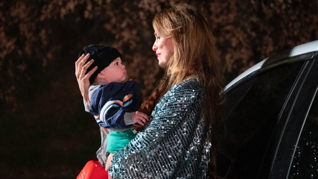 A woman wearing a sparkly, sequined jacket cradles a baby in her arms outside at night, standing beside a car. She gazes at the child with affection, gently cupping his head, while the baby—dressed warmly in a knit hat and jacket—looks back at her with curiosity. The scene is warmly lit, highlighting the tenderness of their connection amid a dark, wooded background.