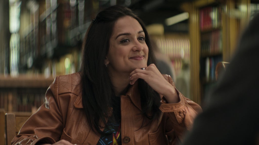 A young woman with long dark hair smiles warmly while seated at a table in what appears to be a library or study room. She wears a tan leather jacket over a patterned shirt and rests her chin on her hand, giving her full attention to someone off-camera. The softly lit background features rows of bookshelves and a scholarly ambiance, suggesting an engaging conversation in an academic setting