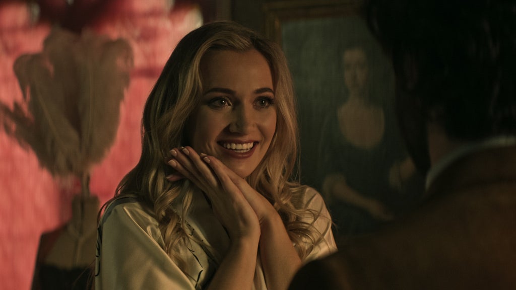 A blonde woman with wavy hair beams with delight, her hands gently pressed together under her chin in a playful, adoring gesture. She wears a silky, light-colored robe and is warmly lit in a cozy, dim room. Behind her hangs a classic-style portrait and soft pink feathers, adding to the scene's rich, vintage ambiance. She appears to be engaging warmly with someone just out of frame.