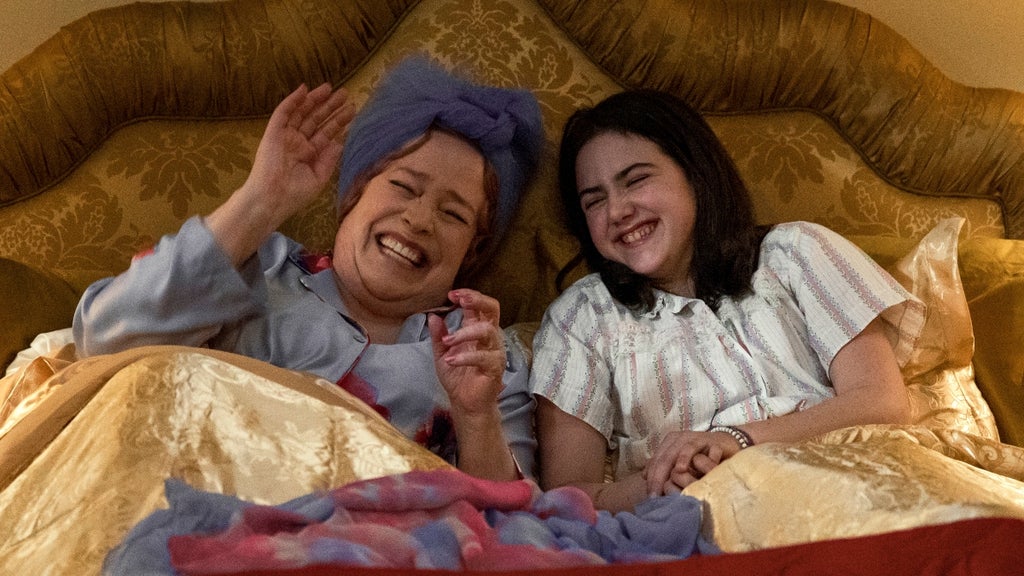 wo women share a joyful moment, laughing together in bed under a golden quilt. The older woman, wearing a lavender head wrap and pajamas, raises her hand mid-laugh, while the younger woman, dressed in a white nightgown with stripes, smiles with her eyes closed.