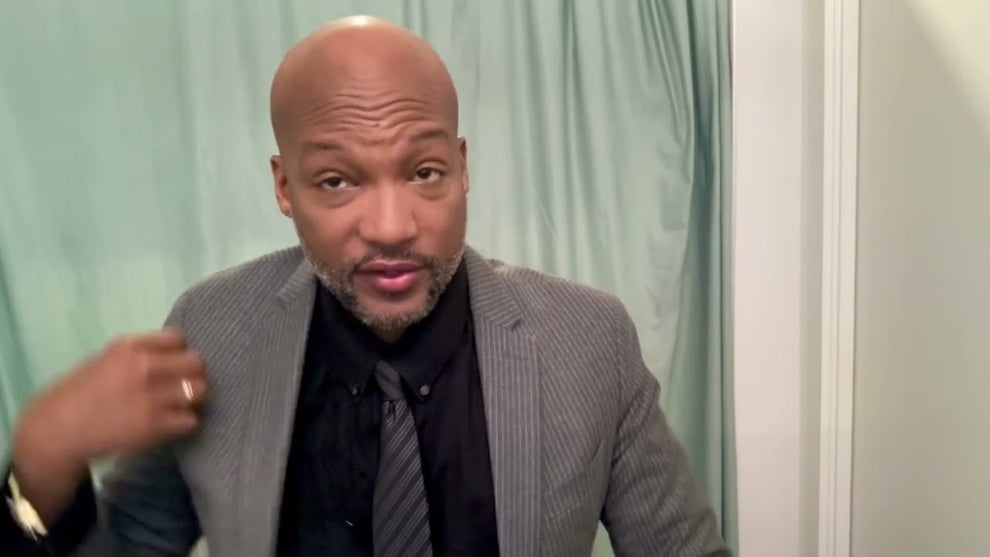 An actor with brown skin, wearing a gray suit, in front of a green backdrop, pulls at his collar.