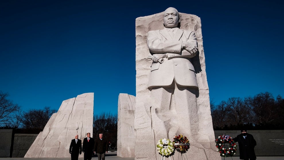 A large white statue of a Black man stands high above a small group of men in suits walking nearby, flowers on a stand, and another nearby man.