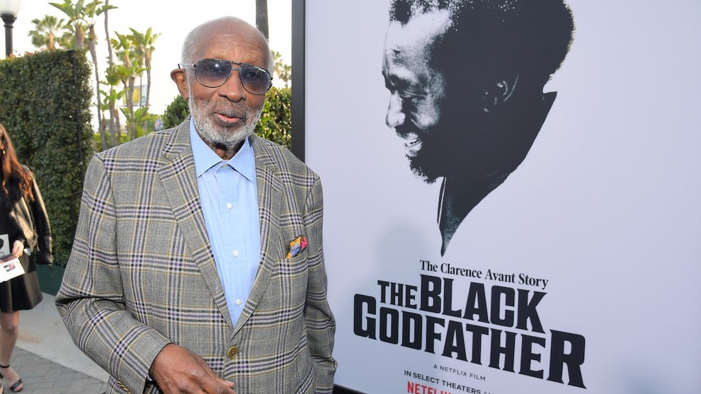 A brown-skinned man wearing sunglasses and a suit stands next to an older photo of his bust on a banner that reads "The Black Godfather"