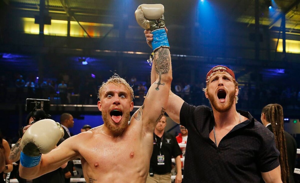 Jake Paul and Logan Paul, two light skin men, pose in a boxing ring. Logan holds his brother Jake's arm up - Jake is wearing boxing gloves and is shirtless, while Logan has a black shirt on. Jake opens his mouth, tongue out, while Logan also has his mouth open and wears a backward baseball cap.