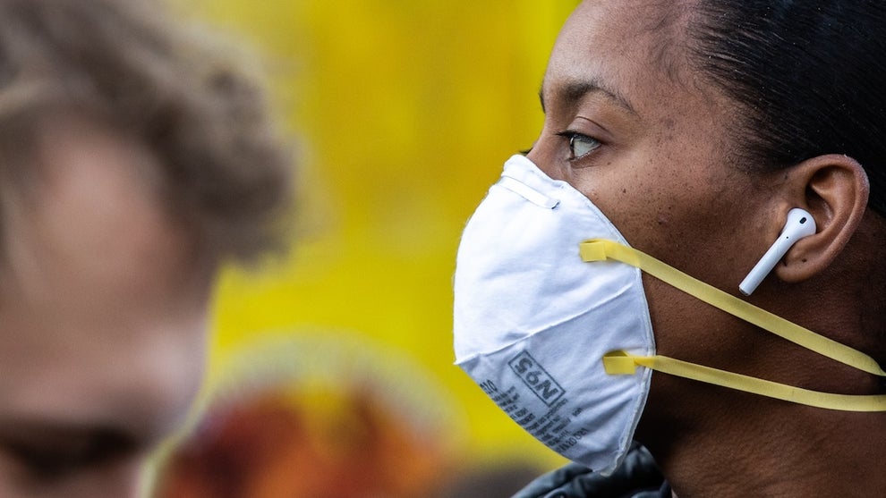 A woman with brown skin wears a mask against a yellow background, a man with light skin walks in the foreground.