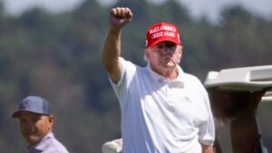 Donald Trump stands outside next to a golf course raising his right fist in the air, wearing a red hat and white golf shirt. Another man stands behind him.