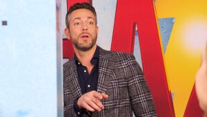 Zachary Levi, a light-skinned man, wears a blazer with button-up shirt with the top buttons unbuttoned, hand up gesturing, in front of a SHAZAM logo