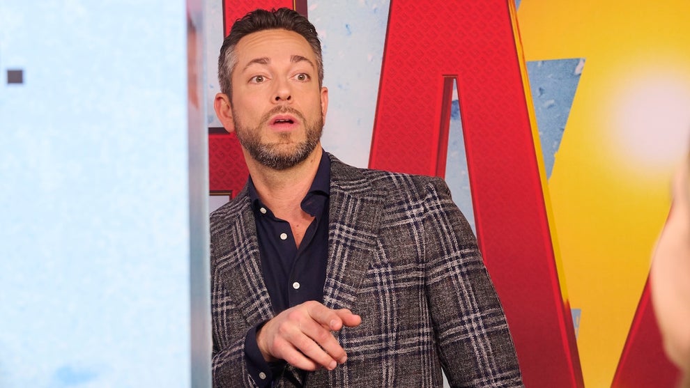 Zachary Levi, a light-skinned man, wears a blazer with button-up shirt with the top buttons unbuttoned, hand up gesturing, in front of a SHAZAM logo