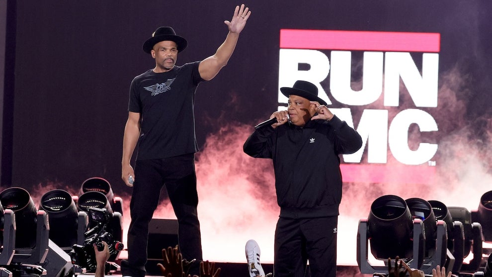 Two men with brown skin stand on stage, the logo for RUN DMC behind them, fans' hands seen in the foreground below with at least one of them holding up an Adidas shoe.
