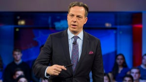 A white man in a suit holds a pen and stands in front of a small audience
