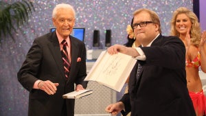 A white man with glasses holds up an autographed book, open to the autographed page - an older white man is behind him to the left of the image, while a young white woman is behind on the right.