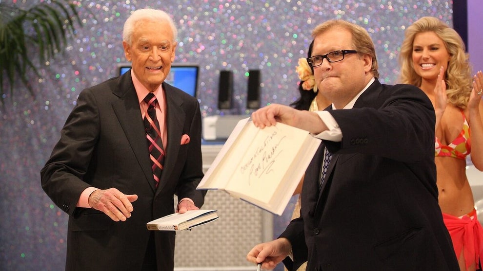 A white man with glasses holds up an autographed book, open to the autographed page - an older white man is behind him to the left of the image, while a young white woman is behind on the right.