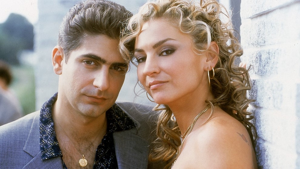 A man and a woman stand next to each other looking at the camera. The man has slicked back black and gray hair while the woman is blonde.