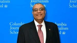 A man with medium-tone brown skin smiles, wears glasses and a suit, in front of a blue background that reads "Columbia Journalism School"