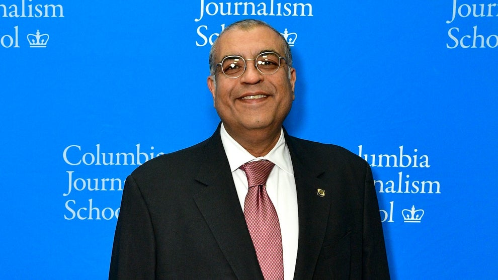 A man with medium-tone brown skin smiles, wears glasses and a suit, in front of a blue background that reads "Columbia Journalism School"