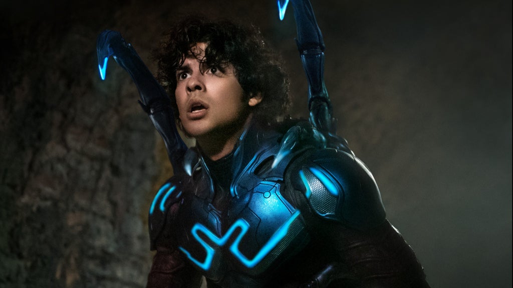 A young Latino man wears a neon blue superhero costume, the blue lit up, with his helmet down, showing his face. There are two antennae sticking up from his shoulders. blue beetle