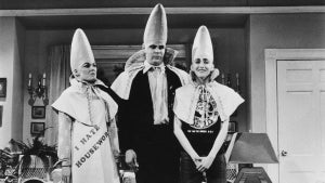 Three people with tall, large, cone-shaped heads (created by prosthetics) on a stage that looks like a living room set.
