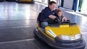 A middle-aged white man drives a yellow bumper car with a female child in his lap, a smile on his face