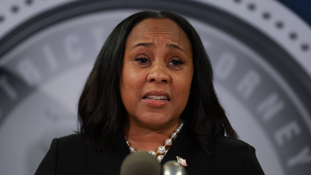 A woman with dark skin looks emotionally toward the camera, in front of a gray background reading DISTRICT ATTORNEY