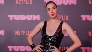 A woman with light-tone skin with her hands on a hip in a shiny black dress, in front of a step-and-repeat reading Netflix Tudum