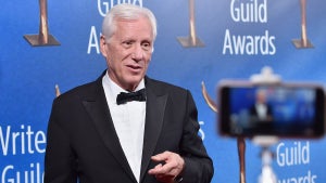 An older white man in a tuxedo is filmed on a smartphone in front of a Writers Guild Awards backdrop