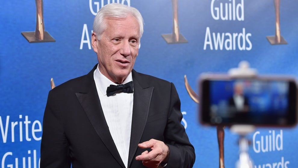 An older white man in a tuxedo is filmed on a smartphone in front of a Writers Guild Awards backdrop