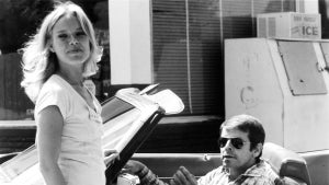 A light-skinned blonde woman in a light T-shirt stands next to a convertible car, a light-skinned man with dark hair and sunglasses sits in the vehicle, which is in front of an outdoor ice machine and windows