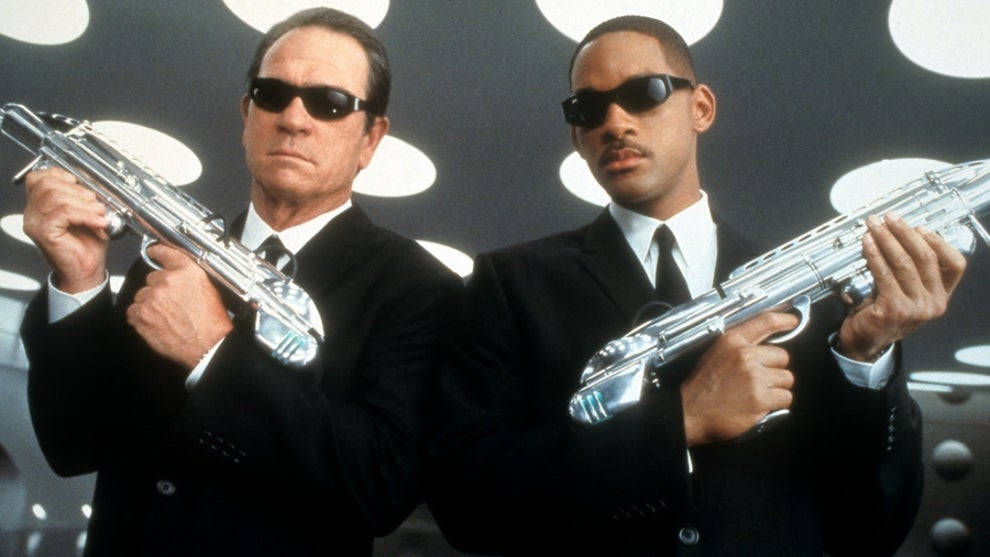 Two men dressed in sleek black suits and dark sunglasses stand confidently with a futuristic cityscape behind them. One holds a silver neuralyzer device, while both exude an air of mystery and authority. Their iconic look suggests they are agents from the "Men in Black" franchise