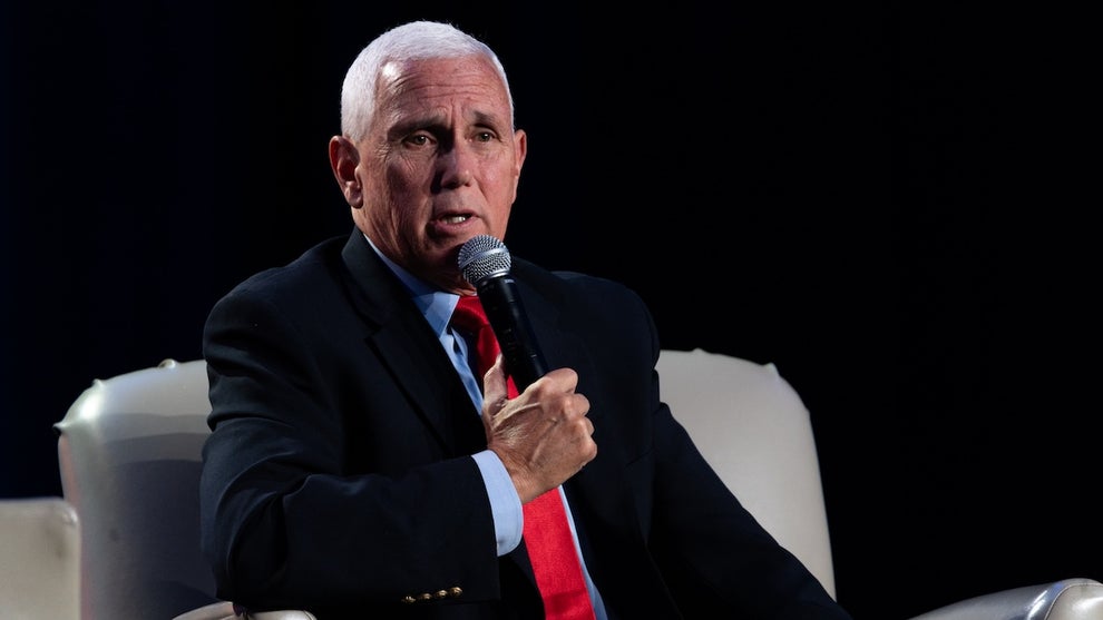 An older white man holds a microphone and wears a suit, sitting down in a sofa