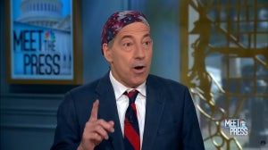 A light-skinned man wearing an American flag head scarf and wearing a suit, raises his finger.