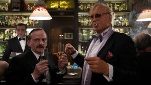 Two older men stand in front of a bar holding drinks