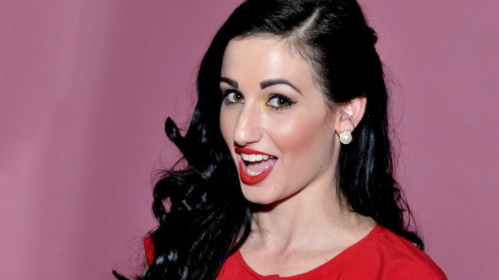 A woman wears a red top and bright red lipstock. She has light-toned skin and long black hair.