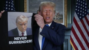 Donald Trump holds up a shirt with his mug shot, with the text "Never Surrender!"