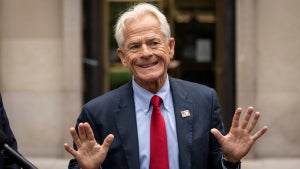 An older white man holds up his hands and looks to be grimacing, wearing a suit and standing outside in front of a building's glass doors.