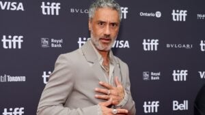 Taika Waititi, a man with medium-tone skin, stands in front of a TIFF step-and-repeat background. He wears a gray suit and crosses his hands across his midsection, with a faux-shocked expression on his face.