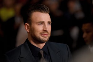 Chris Evans gives just a slight smile, looking to the right, wearing a dark blue suit. He has light skin and dark blonde hair, as well as a bit of a beard.