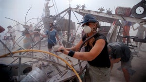 A complex desert vehicle is serviced as people working around it wear face masks.